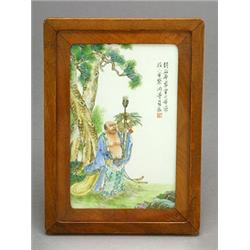 A famille-rose plaque depicting Budai standing under a pine tree, 18cm, framed and glazed (see il...