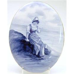 A Royal Doulton oval earthenware plaque painted with a fisher girl seated with basket in blue, 14...