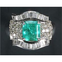 An emerald and diamond shaped-oblong cluster ring, centred with a trap-cut emerald within a surro...