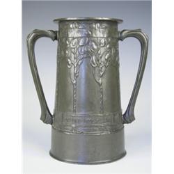 A Tudric pewter two handled tankard, decorated with sinuous stylised trees, and a banded motto...