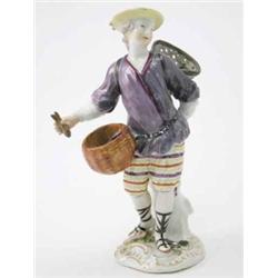 A Meissen figure of a fisherman standing with a net of fish on his back, a circular basket at his...