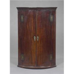 A George III mahogany bow-front hanging corner cupboard, the three shelves enclosed by a pair of...