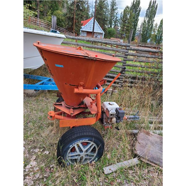 Orange tow-behind broadcast spreader with small gas engine, built-in hopper cover, and trailer hitch