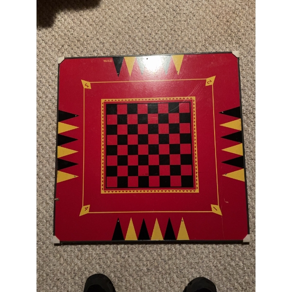 Square wooden game board featuring a central checkerboard in red and black, bordered by backgammon m