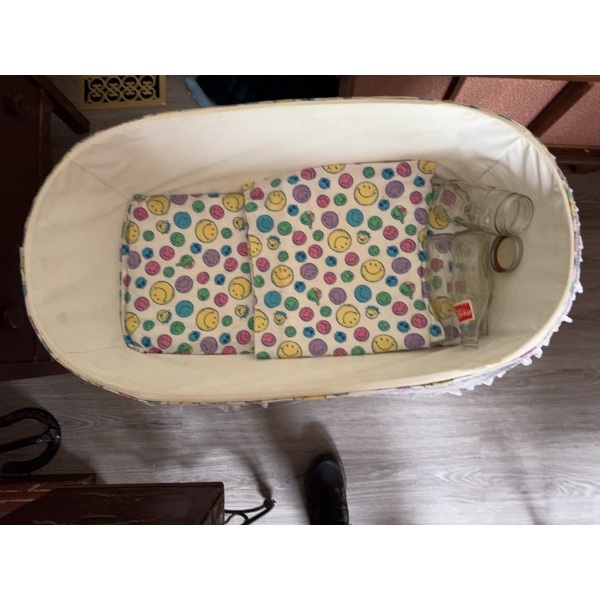 White wicker baby bassinet with folding legs, lace trim, fitted colorful smiley face fabric, and thr