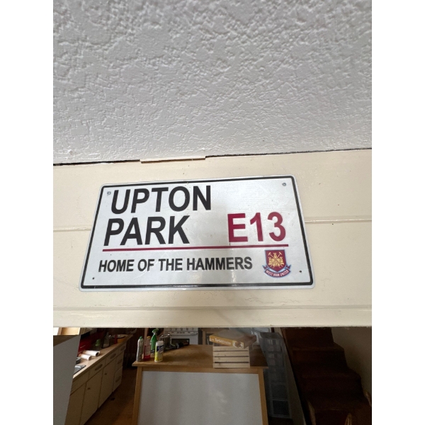 A decorative metal sign plaque featuring the text "UPTON PARK E13 HOME OF THE HAMMERS" and the West 